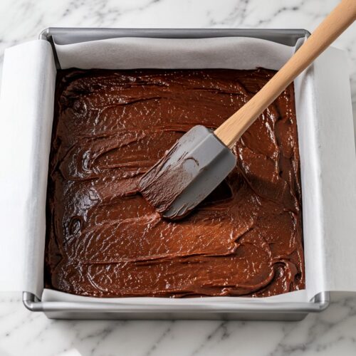 A silicone spatula spreading dark chocolate brownie batter in a parchment-lined baking pan.