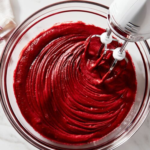 Electric hand mixer blending red velvet batter in a clear glass bowl.