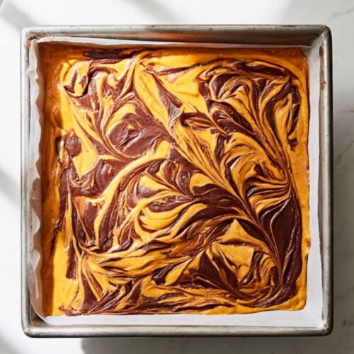 A square pan lined with parchment shows a vibrant, swirled pattern of chocolate and pumpkin batters.