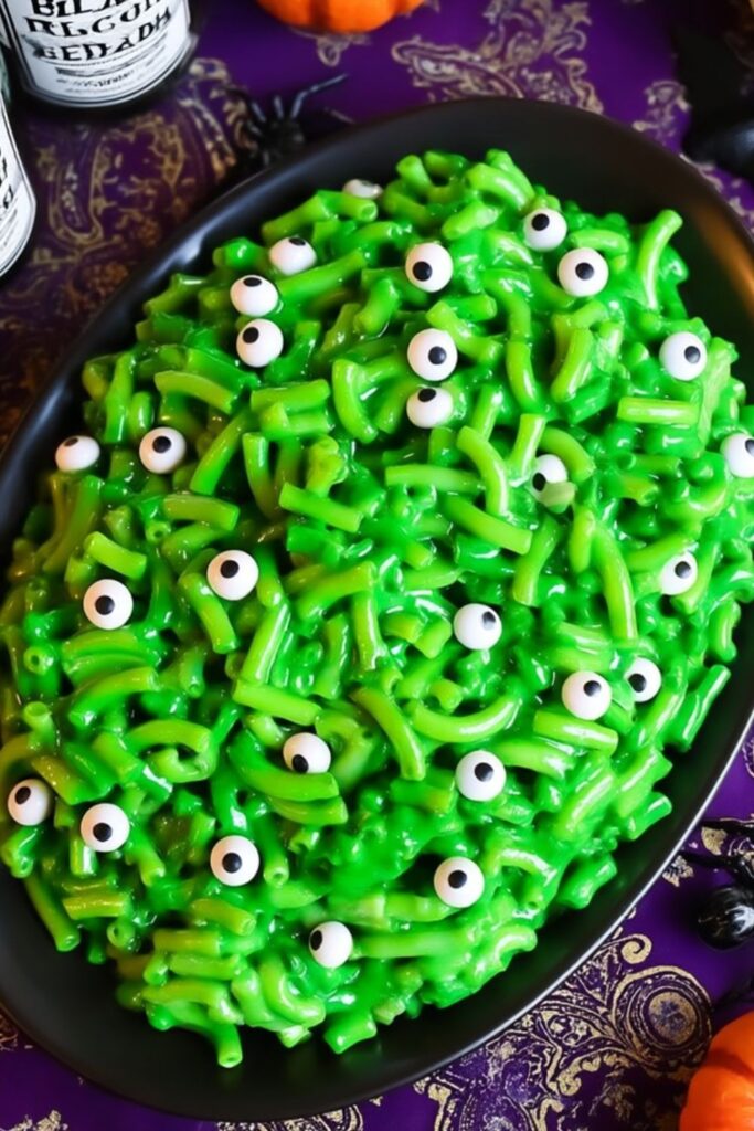 Bright green mac and cheese topped with edible candy eyes on a black platter.