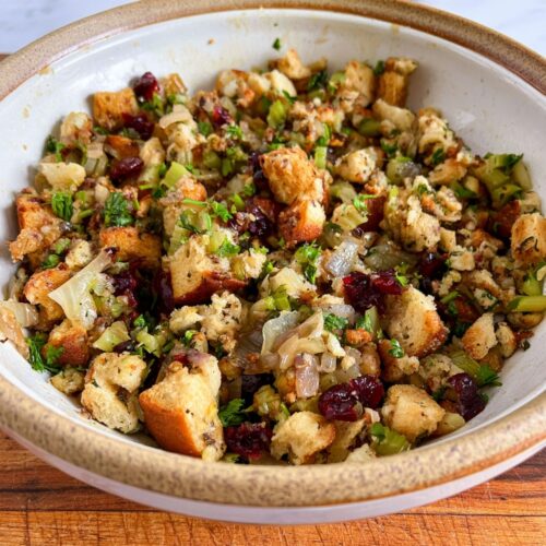 A ceramic bowl filled with a savory mixture of toasted bread cubes, cranberries, onions, celery, and herbs.