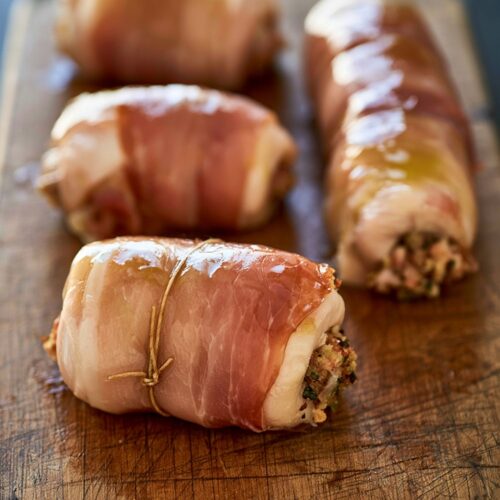Stuffed chicken roulades wrapped in prosciutto and tied with twine on a wooden board.