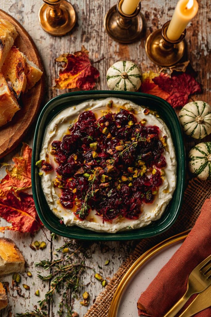 A green bowl of whipped feta topped with cranberry sauce and pistachios next to sliced bread.
